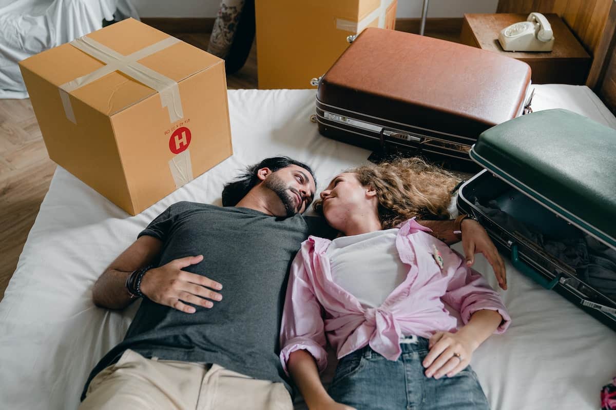 From above of young ethnic bearded man and woman with curly hair looking at each other and lying on bed among suitcases and cardboard boxes with stuff while moving in new apartment together