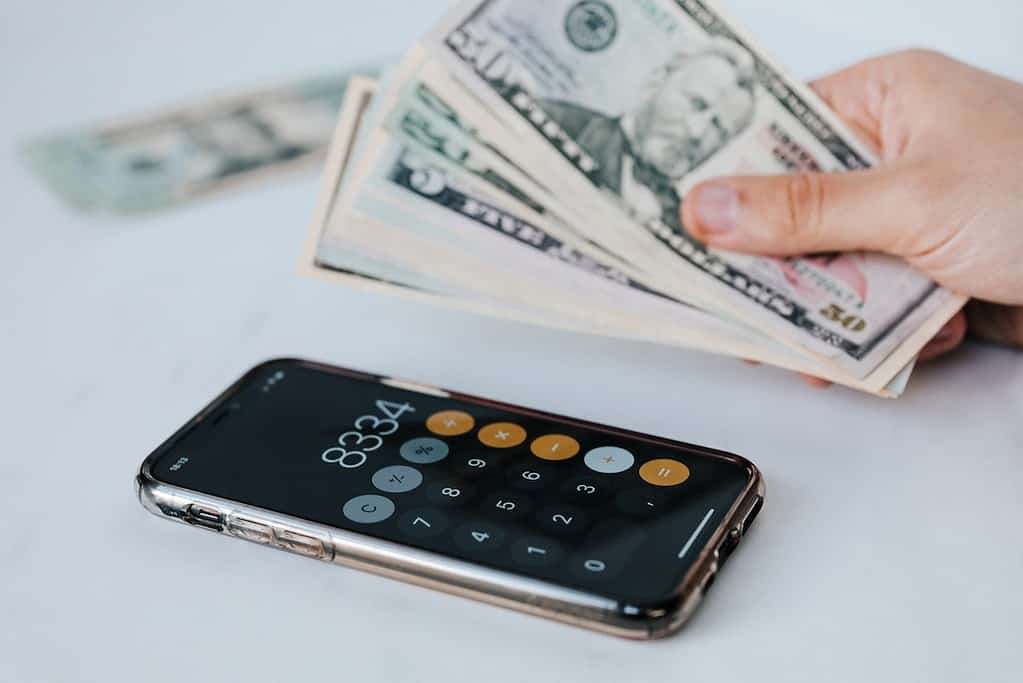 Calculator application on mobile phone screen on white surface  person holding pile of dollars